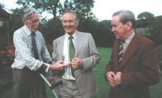 Donald Sinclair, Alf Wight, and Brian Sinclair (left to right) are smiling. Donald is wearing checkered long sleeves, a blue tie, and brown pants, Alf is wearing a gray coat over white long sleeves and a green tie while Brian is wearing a brown coat over gray long sleeves and a striped brown and white tie.
