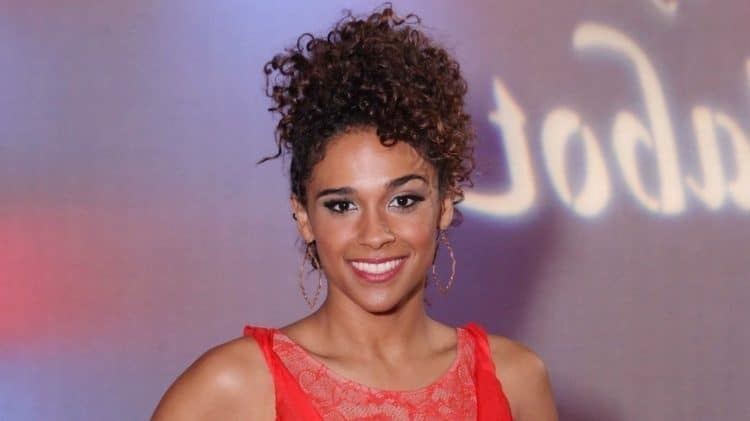 Briana Henry smiling while her curly hair is tied up and she is  wearing a red sleeveless blouse and hoop earrings
