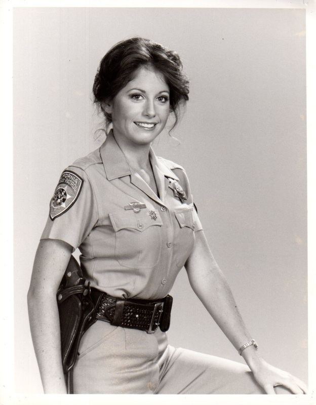 Brianne Leary is smiling, has wavy hair, wearing a wristwatch on her left hand, a gray police uniform with a police badge on her left chest, a pin on her right chest, and a police shoulder badge on her right shoulder, a black police duty belt with a gun on her right waist.