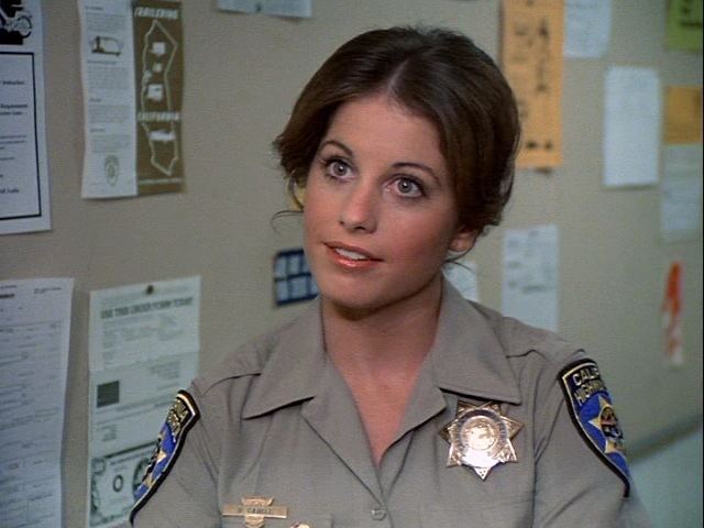 Brianne Leary is smiling, has brown wavy hair, and a bulletin board at her back with papers, she is wearing a gray police uniform with a police badge on her left chest, a pin on her right, and a police shoulder badge on both of her shoulders.