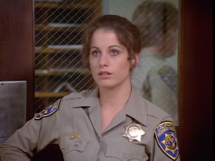 Brianne Leary is serious and has brown wavy hair, she is standing in front of the door, mouth half opened, and she is wearing a gray police uniform with a police badge on her left chest, a pin on right, and a police shoulder badge on both of her shoulders.