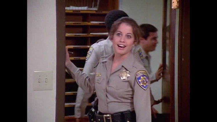 Brianne Leary is smiling, has brown wavy hair, right hand on the wall, behind her are two policemen, she is wearing a gray police uniform with a police badge on her left chest, a pin on her right chest, a police shoulder badge on her left shoulder, and a black police duty belt with a gun on her right waist.