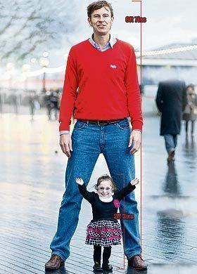 Bridgette Jordan is smiling while standing in front of a man comparing their heights. Bridgette is wearing eyeglasses, a black and white top, a multi-colored skirt, and black boots while the man is wearing a red sweatshirt, blue denim pants, and brown shoes.