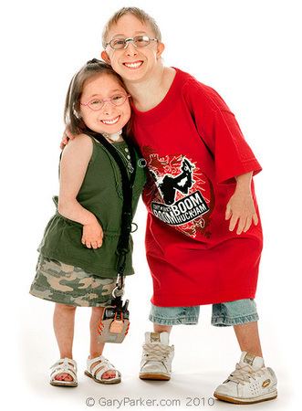 Bridgette and Brad Jordan are smiling. Bridgette is wearing eyeglasses and a lengthy necklace, wearing a green top, a camouflage skirt, and white sandals while Brad is wearing eyeglasses, a red shirt with prints, blue shorts and white shoes.