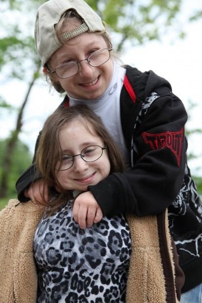 Bridgette and Brad Jordan are smiling while Brad is hugging Bridgette. Bridgette is wearing eyeglasses, wearing a black and white top, and a brown scarf while Brad is wearing eyeglasses, a brown cap, a multi-colored jacket over a white shirt, and blue jeans.