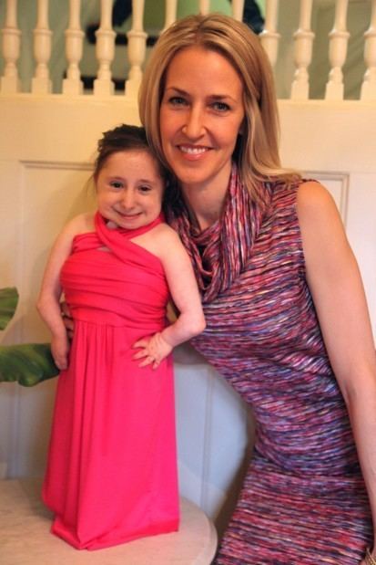 Bridgette Jordan and Amy Johnson are both smiling. Bridgette is wearing a pink halter dress while Amy, holding her is a KayOss Designs' fashion designer, has blonde hair, wearing a multi-colored sleeveless dress.