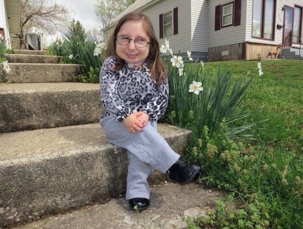 Bridgette Jordan is smiling with houses in the background while sitting with crossed legs beside white flowers, wearing eyeglasses, a black and white long sleeve top, blue jeans, and black shoes.