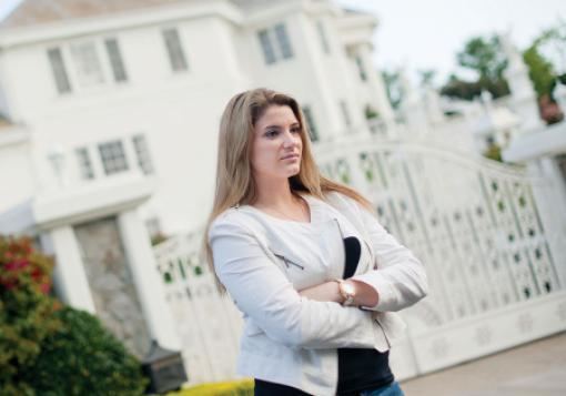 Brittany Koper is serious, hands crossed standing in front of a green bush and a white mansion with white large gate, has long blond hair wearing a silver watch, a block top under a white coat and denim pants.
