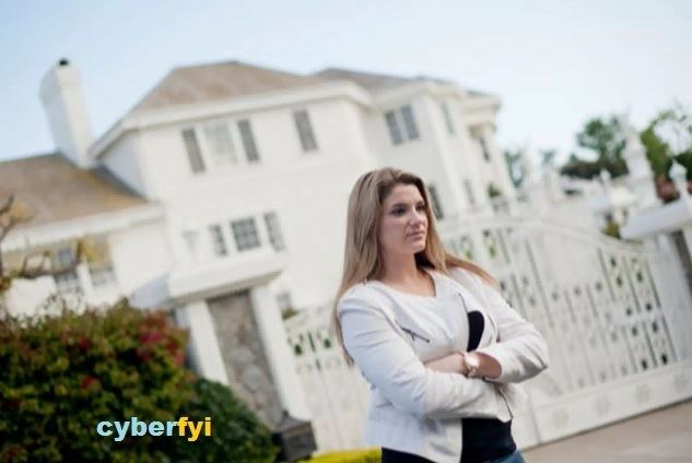 Brittany Koper is serious, hands crossed standing in front of a green bush and a white mansion with white large gate, has long blond hair wearing a silver watch, a block top under a white coat and denim pants. At the let bottom is a word “cyberfyi”