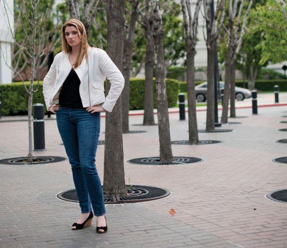 In a park with trees with decorative streetscapes and brick floors, Brittany Koper is serious, standing with her hands on her waist, has long blond hair wearing a black shirt under a white coat and denim pants with a black high heels.