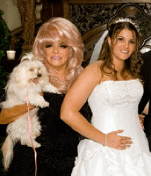 In a room with large antique brown walls at the back, from left, Jan Crouch is smiling, standing holding a white Maltese dog with pink lace, has Pink hair wearing a gold necklace and a black dress, at the right Brittany Koper is smiling, standing, with her right hand in her waist, has curly brown hair wearing a white wedding veil and a white wedding gown.