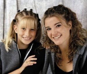 In a room with a gray curtain, from left, Carra Crouch is smiling, sitting, with her right hand on Brittany’s shoulder, has braided blond hair with bangs, wearing a black shirt under a gray jacket, at the left, Brittany Koper is smiling, sitting, has brown curly hair wearing a black shirt under a gray jacket with black sleeves.
