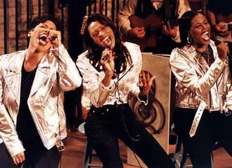 Members of Brownstone, Mimi Doby, Nicci Gilbert, and Charmayne Maxwell are singing on the stage, holding microphones, wearing white jackets and black pants.