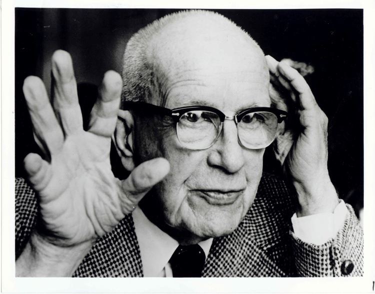 Buckminster Fuller's hand on his head while wearing eyeglasses, checkered coat, long sleeves, and necktie