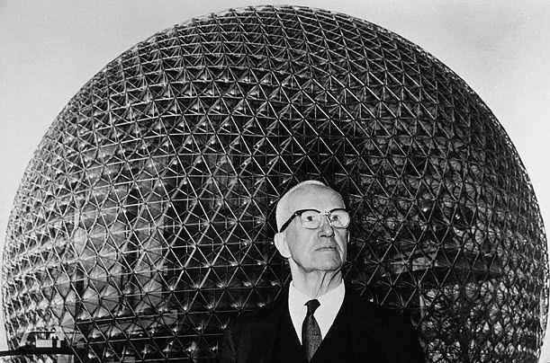 Buckminster Fuller standing in front of his geodesic dome while wearing eyeglasses, coat, long sleeves, and necktie