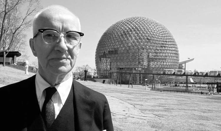 Buckminster Fuller smiling and wearing a coat, long sleeves, necktie, and eyeglasses
