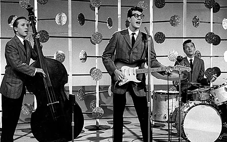 Buddy Holly, center, singing while playing guitar with Drummer Jerry Allison, and Bassist Joe B. Mauldin beside him and they are all wearing striped coats, long sleeves, black neckties, and black pants