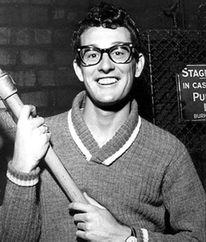 Buddy Holly with a big smile while holding a stick and wearing eyeglasses, sweater with collar, and wristwatch