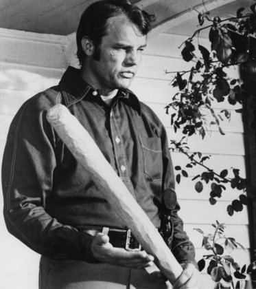 Buford Pusser is serious, has black hair, both hands holding a baseball bat, and wears black long sleeves, gray pants, and a black belt.