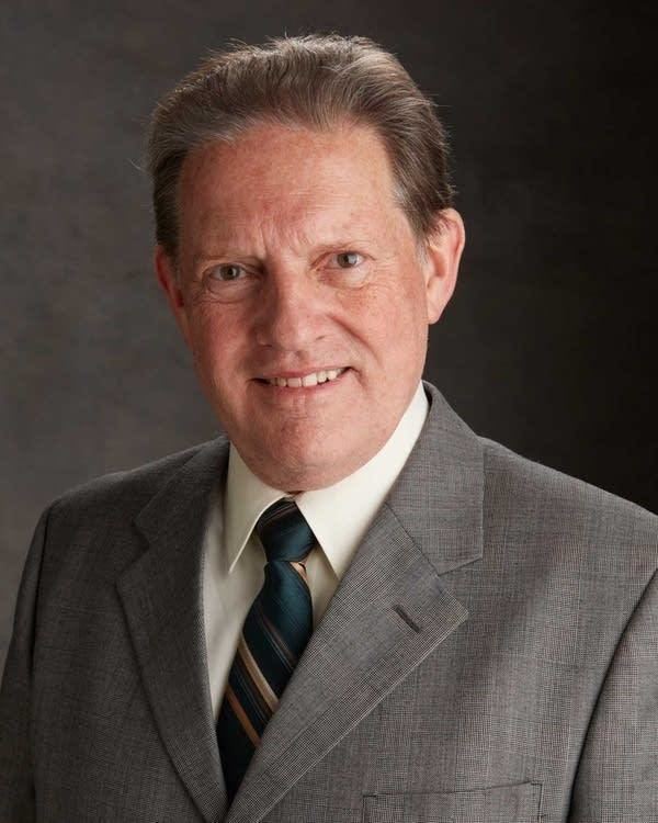 Byron David Smith smiling while wearing gray coat, cream long sleeves and striped neck tie