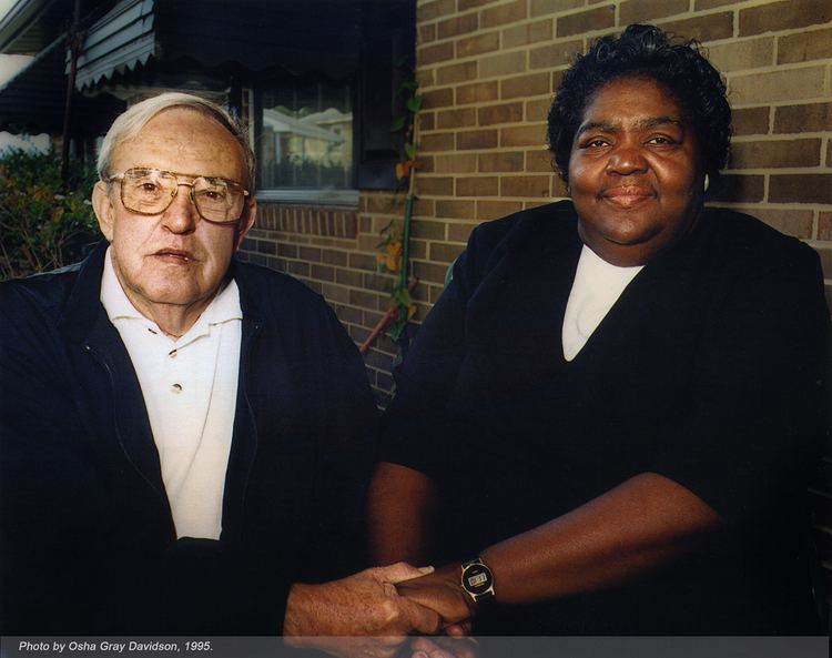 C. P. Ellis wearing eyeglasses, a white shirt, and a black coat while holding Ann Atwater wearing a black and white shirt.