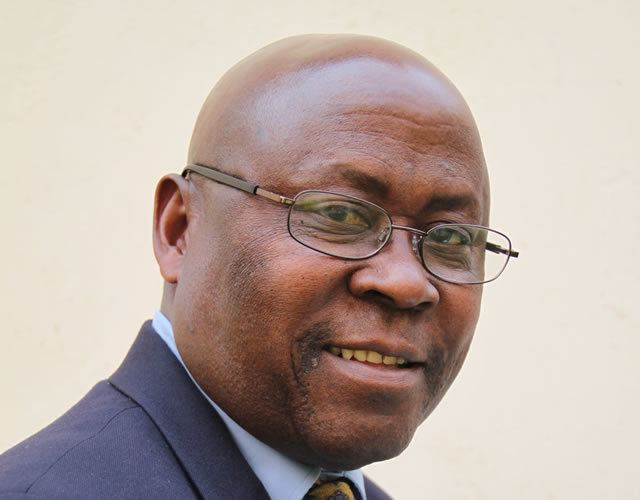 Cain Mathema smiling while wearing a dark blue coat, light blue long sleeves, yellow necktie, and eyeglasses