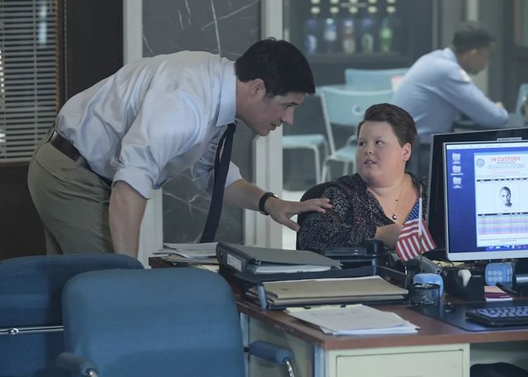 Rich Sommer and Calle Walton talking to each other in the office with computers, files, and an American flag on the desk, and another person in the other room behind Calle Walton and Rich is wearing light blue long sleeves while Calle is wearing a black blouse and a necklace