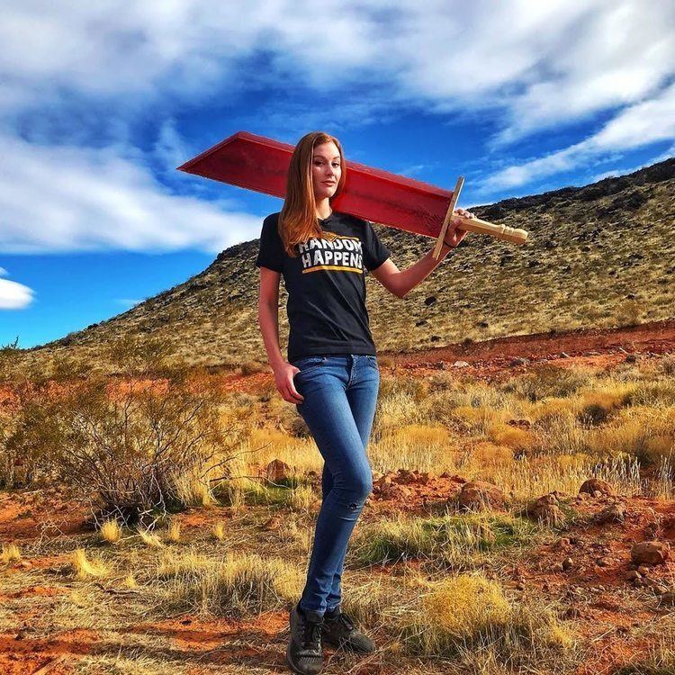 Calli Gade smiling while carrying a big red sword, with blonde hair, wearing a black shirt, blue jeans, and black shoes.