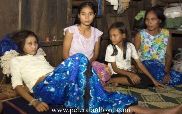 Rochom P’ngieng is lying on the bed and her mother, Rochom Soy and two other women are beside her. P’ngieng is wearing a cream blouse and a blue skirt