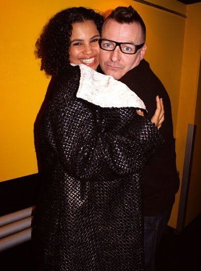 Cameron McVey's wife, Neneh Cherry ( left) is smiling, has black curly hair, wearing a long black suit, hugging her husband Cameron McVey (right) is serious and has a mustache and black hair, wearing his eyeglasses, a black jacket, and black pants.