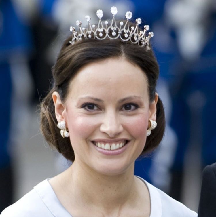 Carina Axelsson is happy, has long black hair tied up, is wearing a silver with diamond crown, silver with diamond earrings, and a white dress.
