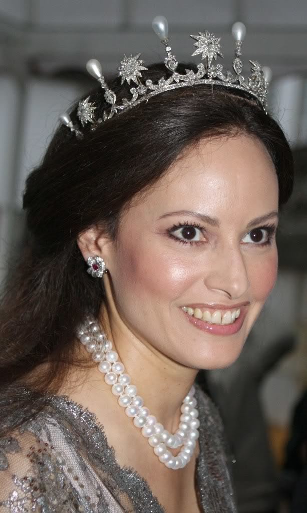 Carina Axelsson is happy, has long black hair, wearing a silver with diamonds and pearls crown, silver flower earring, white pearl necklace, a white slip under a silver see through gown.
