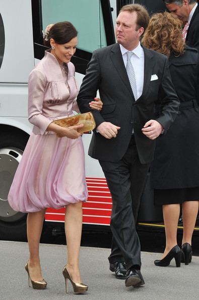 On the left Carina Axelsson is happy, walking with Prince Gustav behind them is a white bus and a woman (right), her right hand holding a small yellow-brown bag while left hand holding Prince Gustav left arm, has long black hair tied behind with a white hair clip, wearing silver earrings, a ruby ring and a pink dress with gold high heels, In the middle, Gustav, 7th Prince of Sayn-Wittgenstein-Berleburg is serious, looking at his side while walking beside Carina Axelsson, wearing a white polo with white bowtie, a watch under the black tuxedo and black shoes. At the right, a woman at her back, has brown curly hair wearing a black coat, skirt and black high heels. Inside the bus is a man, looking down, with gray hair wearing a white polo with red necktie and black coat.