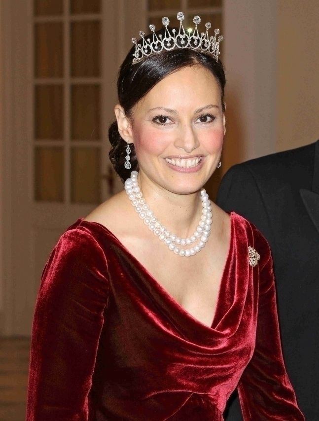 Carina Axelsson is happy, has long black hair tied at the back, wearing a silver with diamond crown, silver with diamond dangle earring, white pearl necklace, and a red long-sleeve dress with diamond brooches below the left shoulder.