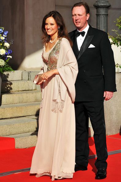On the left Carina Axelsson is happy, standing on the red carpet with blue and white flowers on the left, with her right hand holding a small brown bag, has long black hair, wearing silver earrings, a ruby ring, pearl necklace and a beige gold chest design gown. At the right, Gustav, 7th Prince of Sayn-Wittgenstein-Berleburg is serious, standing behind Carina Axelsson, wearing a white polo with black bowtie under the black tuxedo and black shoes.