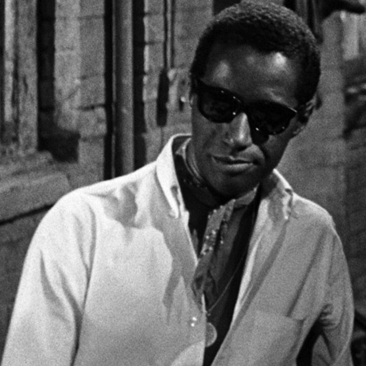 Carl Lee with a tight-lipped smile while wearing sunglasses, ribbon scarf, and long sleeves in a scene from the 1961 film, The Connection