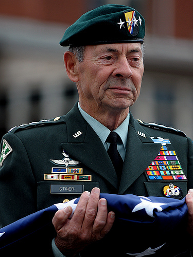 Carl Stiner is serious, standing while holding a folded flag on top of his hands a four Star General, has faded black hair wearing a US Army Ranger Uniform with white top,  black tie, with badges on his chest.
