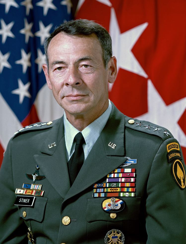 Carl Stiner is serious, behind is the USA flag, he is a four Star General, has faded black hair wearing a US Army Ranger Uniform with white top,  black tie, with badges on his chest and broided ranger airborne title on arms.