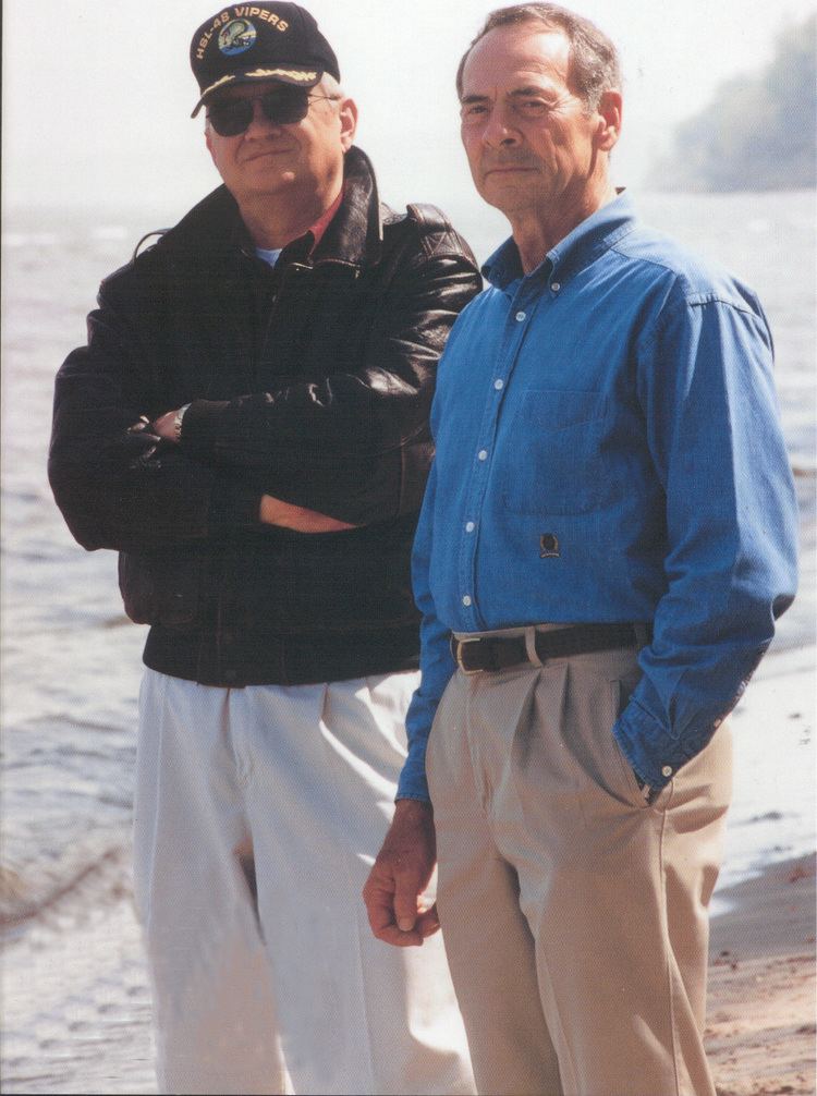 Tom Clancy (left) is smiling standing next to Carl Stiner (right) has faded hair wearing shades and an army cap, leather jacket and white pants, Carl Stiner (right) is serious, left hand inside his pocket he is a four Star General, has faded black hair wearing a blue top black belt and a khaki pants.