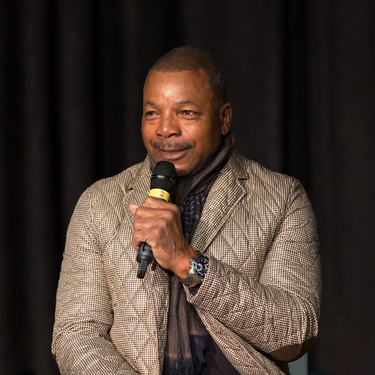 Carl Weathers speaking through a microphone and wearing a gray coat and violet scarf