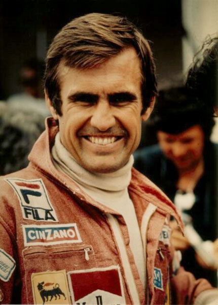 Carlos Reutemann smiling while wearing a red racing suit and white turtle neck long sleeves