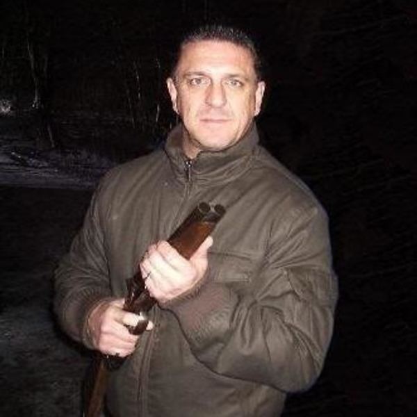 Carlton Leach with a serious face, wearing a brown jacket while holding a gun.