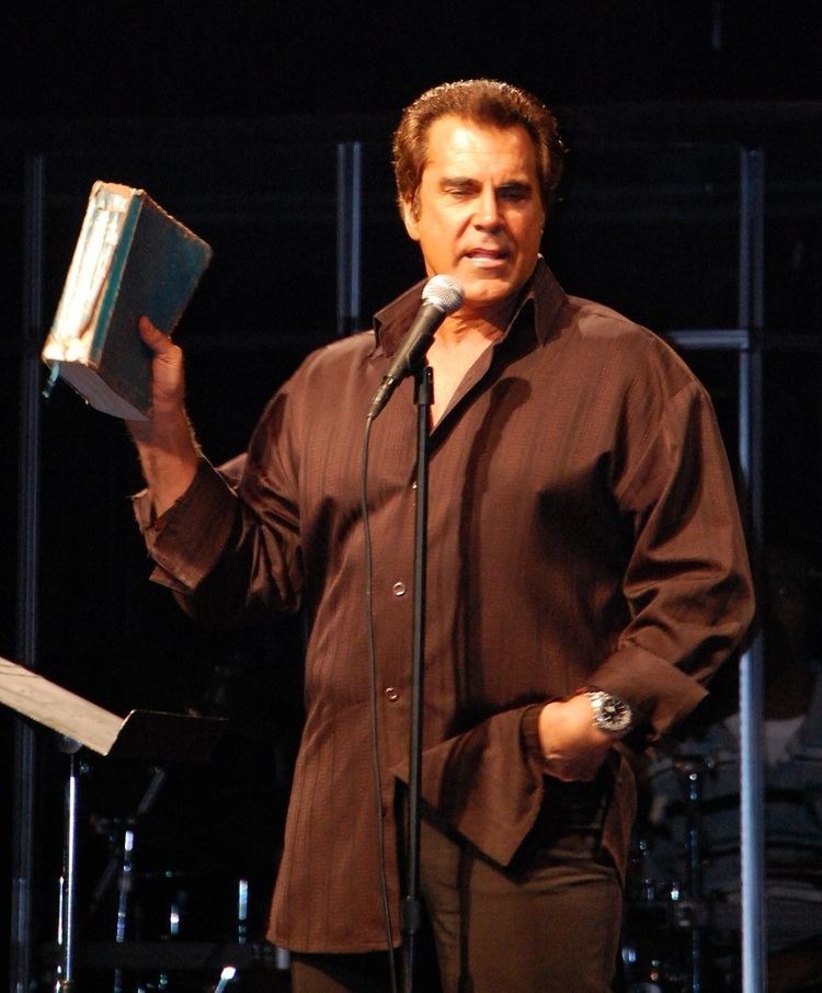 Carman giving a speech while holding a book and wearing a brown long sleeve, brown pants, and a wristwatch