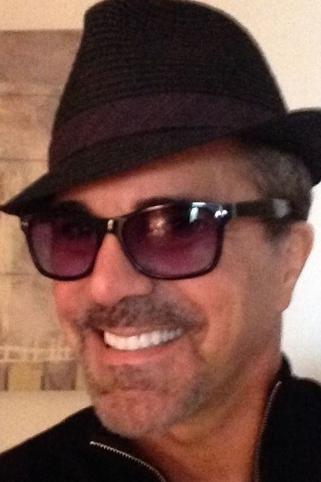 Carman smiling with mustache and beard while wearing a black hat, sunglasses, and black polo
