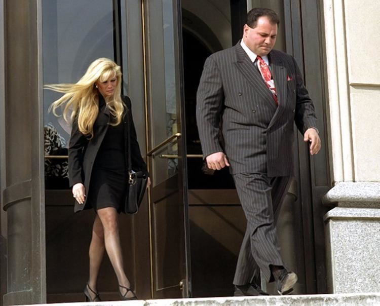 Carmine Agnello wearing black striped coat paired with black striped pants, white long sleeves and red neck tied and a woman at his back wearing black coat and black skirt