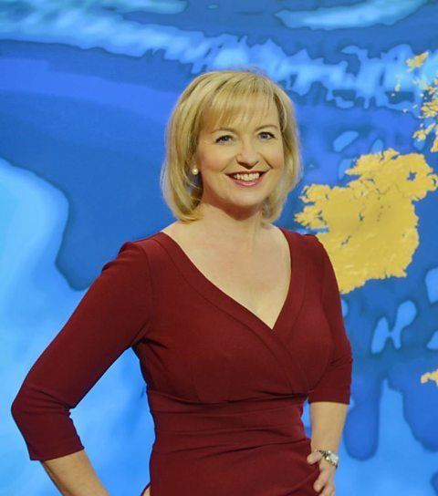 Carol Kirkwood's hands on her hips while wearing a maroon dress