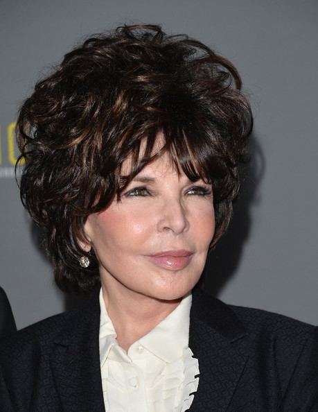 Carole Bayer Sager with a tight-lipped smile while looking afar and wearing a black coat, white blouse, and earrings