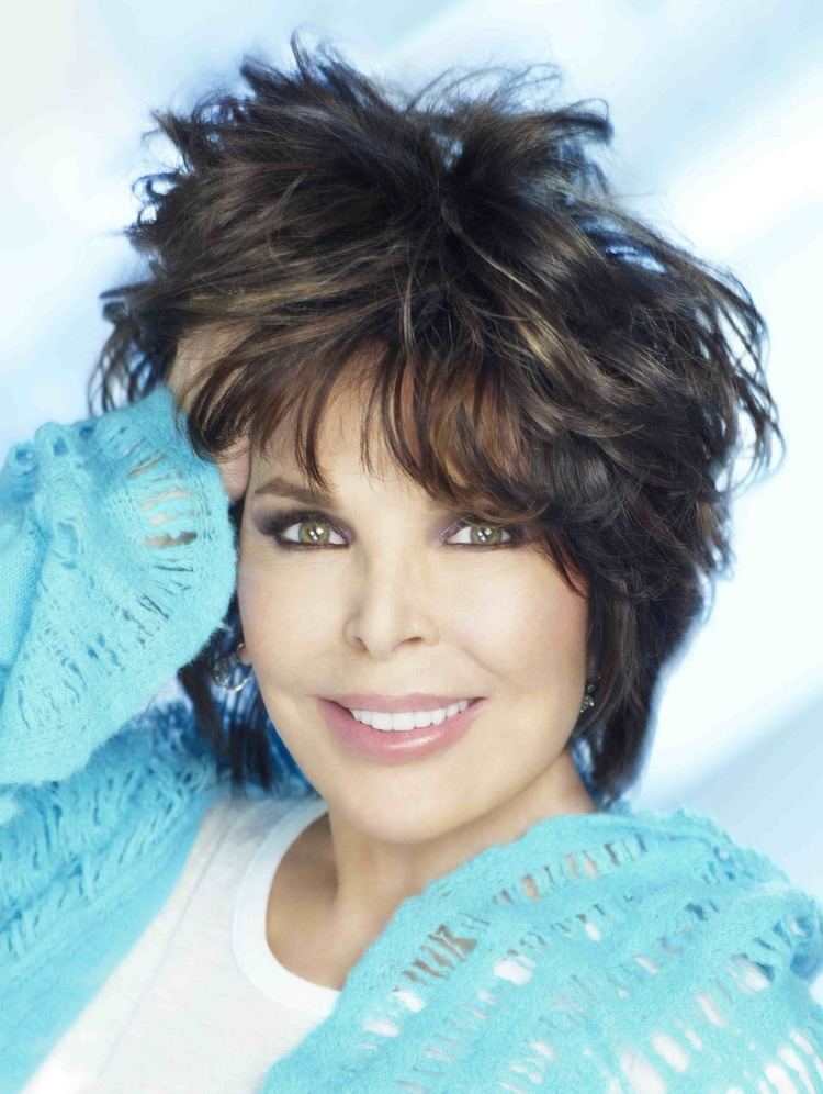 Carole Bayer Sager smiling while hand on her head and wearing a blue blazer and white blouse