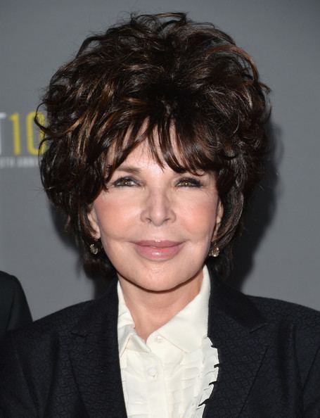 Carole Bayer Sager in her tight-lipped smile and short hair with layers while wearing a black coat, white blouse, and earrings