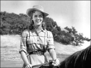 Carolyn Craig smiling while riding a horse, wearing a hat, a checkered blouse, and a belt.